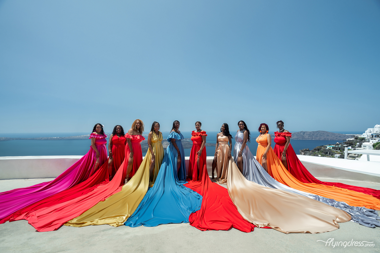A vibrant group of women in flowing, colorful flying dresses poses on a Santorini terrace with the stunning Aegean Sea as their backdrop.